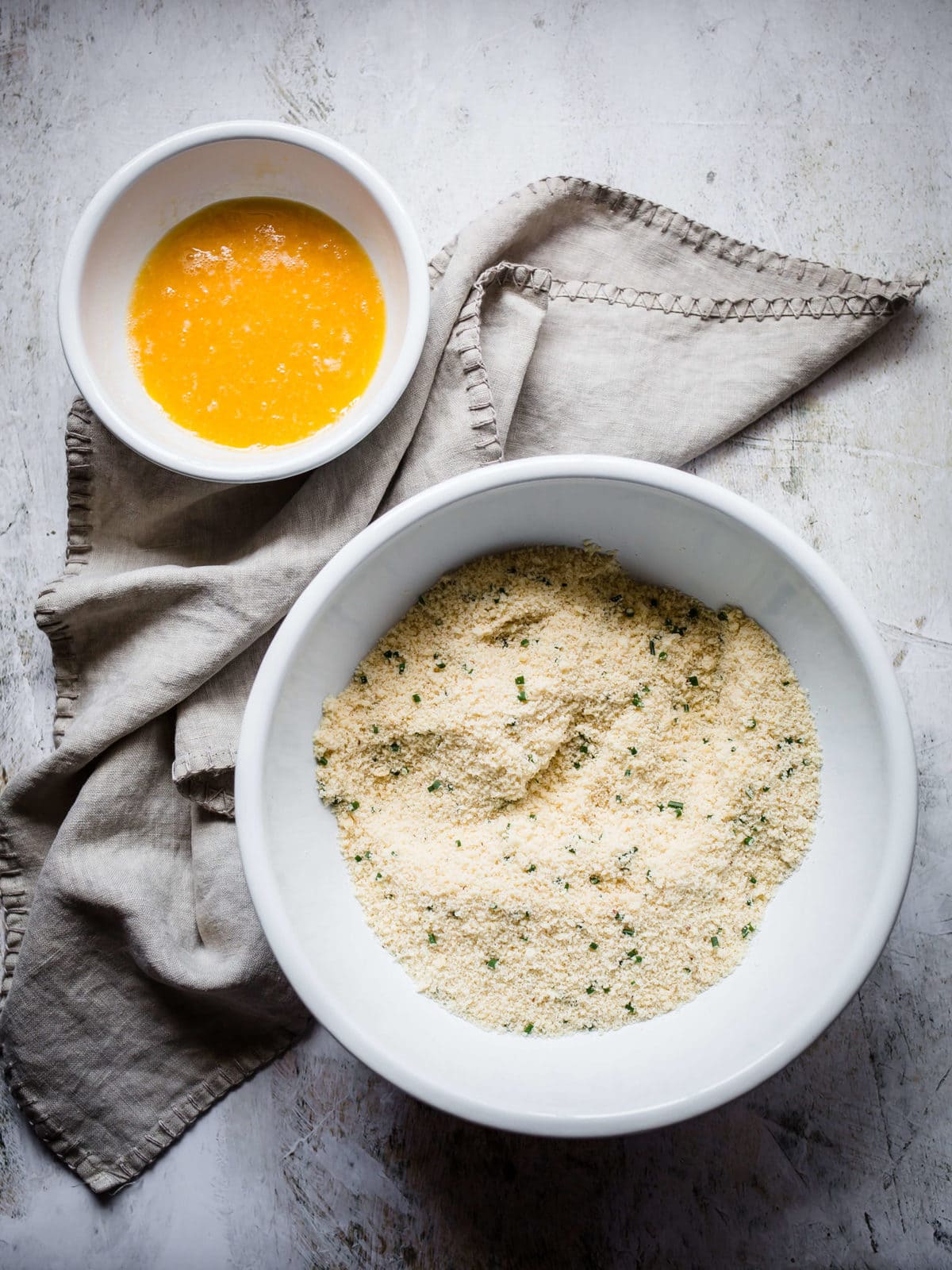 A mixing bowl with almond flour, grated parmesan cheese, garlic powder and chopped fresh chives, with a bowl of egg wash next to it.