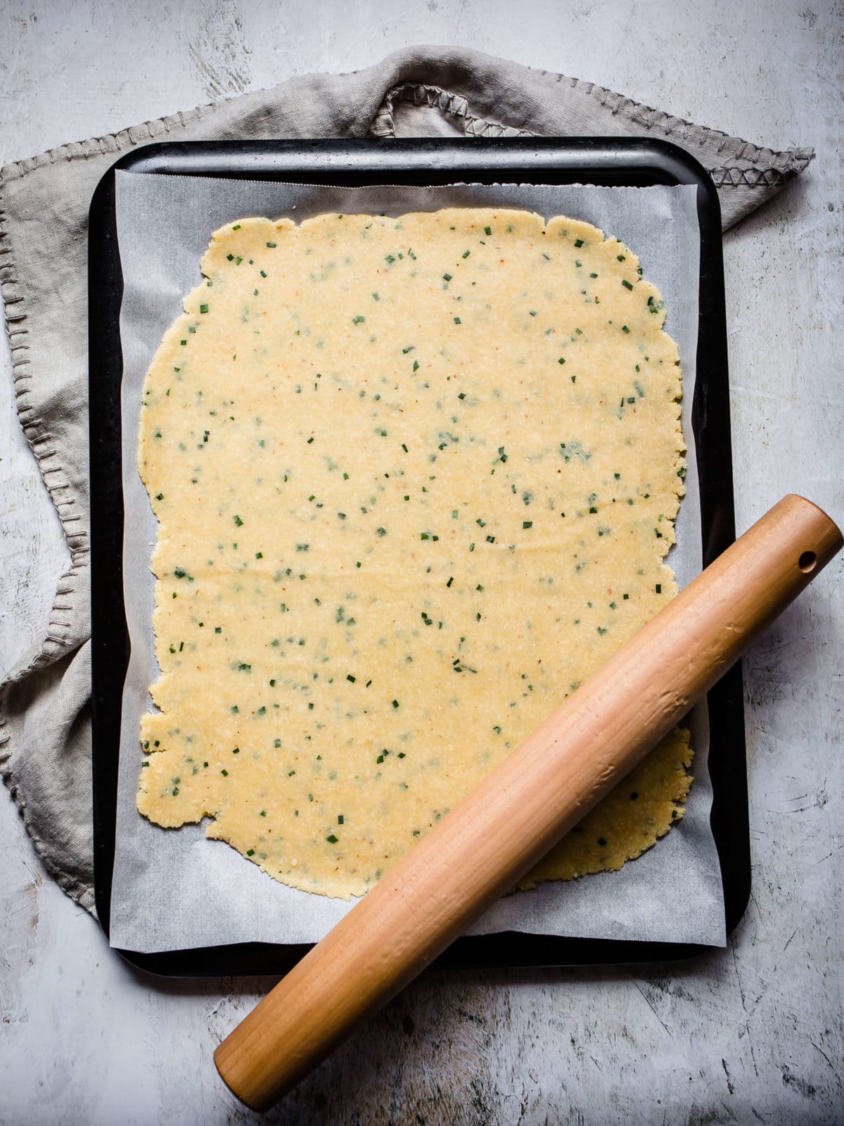 Keto cracker dough with chives rolled out on parchment paper on a baking sheet.