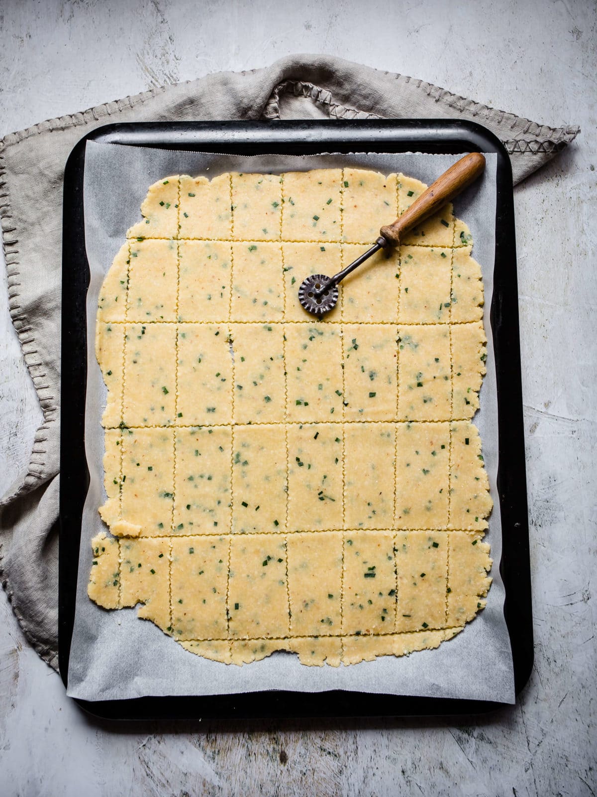 Keto cracker dough with chives rolled out on parchment paper on a baking sheet and cut into individual crackers.