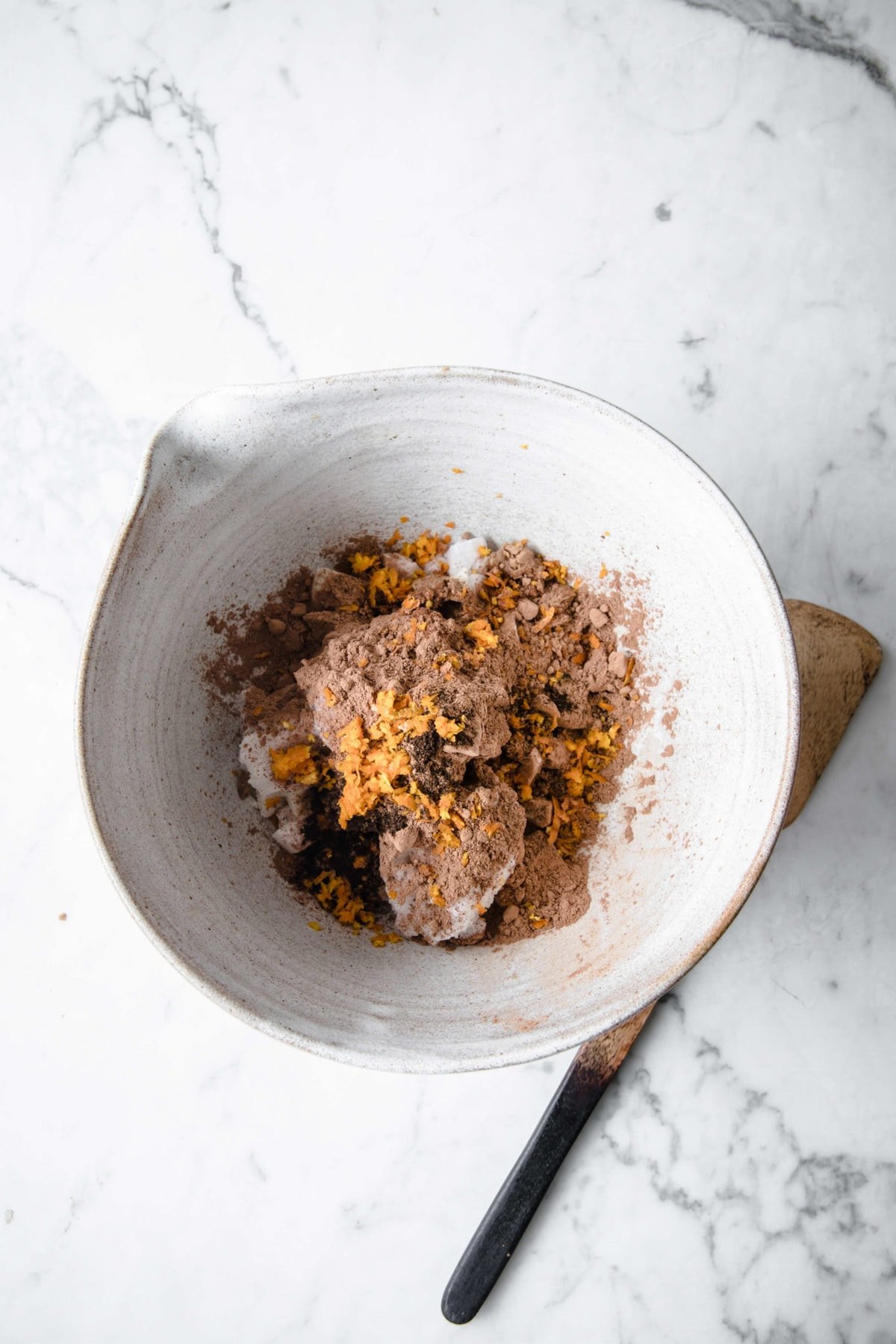 a ceramic mixing bowl with a wooden spoon with coconut oil, orange zest, and unsweetened cocoa powder