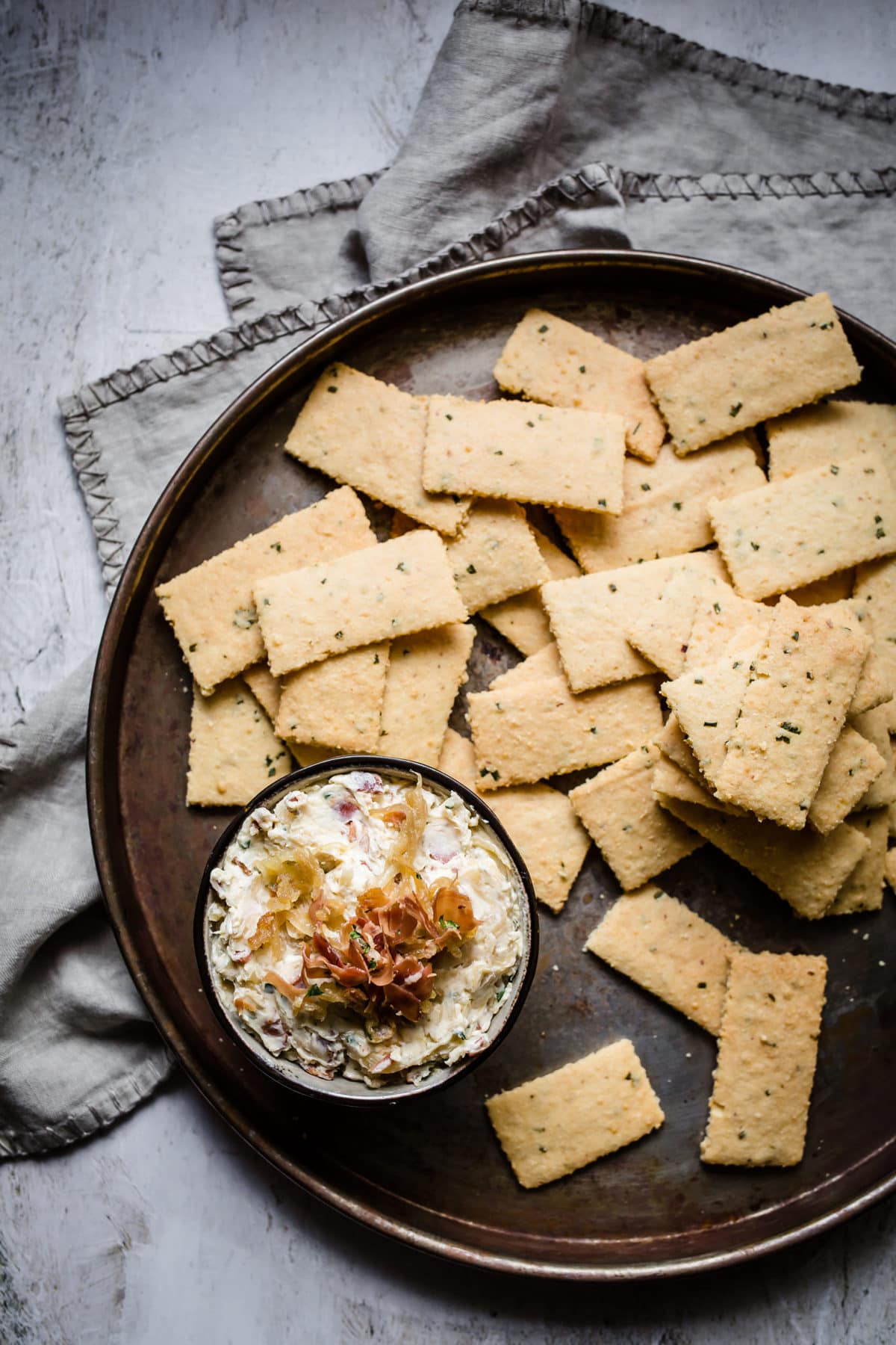 A pizza pan full of garlic parmesan keto crackers, served with caramelized onion and bacon dip with a gray towel next to it.