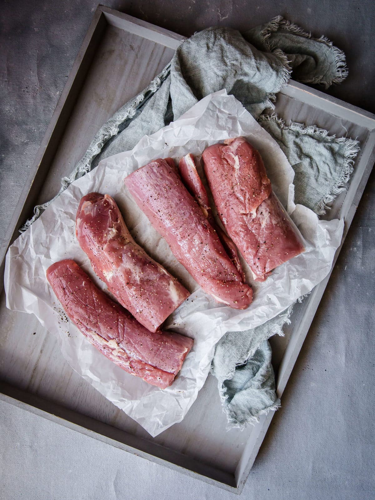 four raw pork tenderloins seasoned with salt and pepper, lined on parchment paper