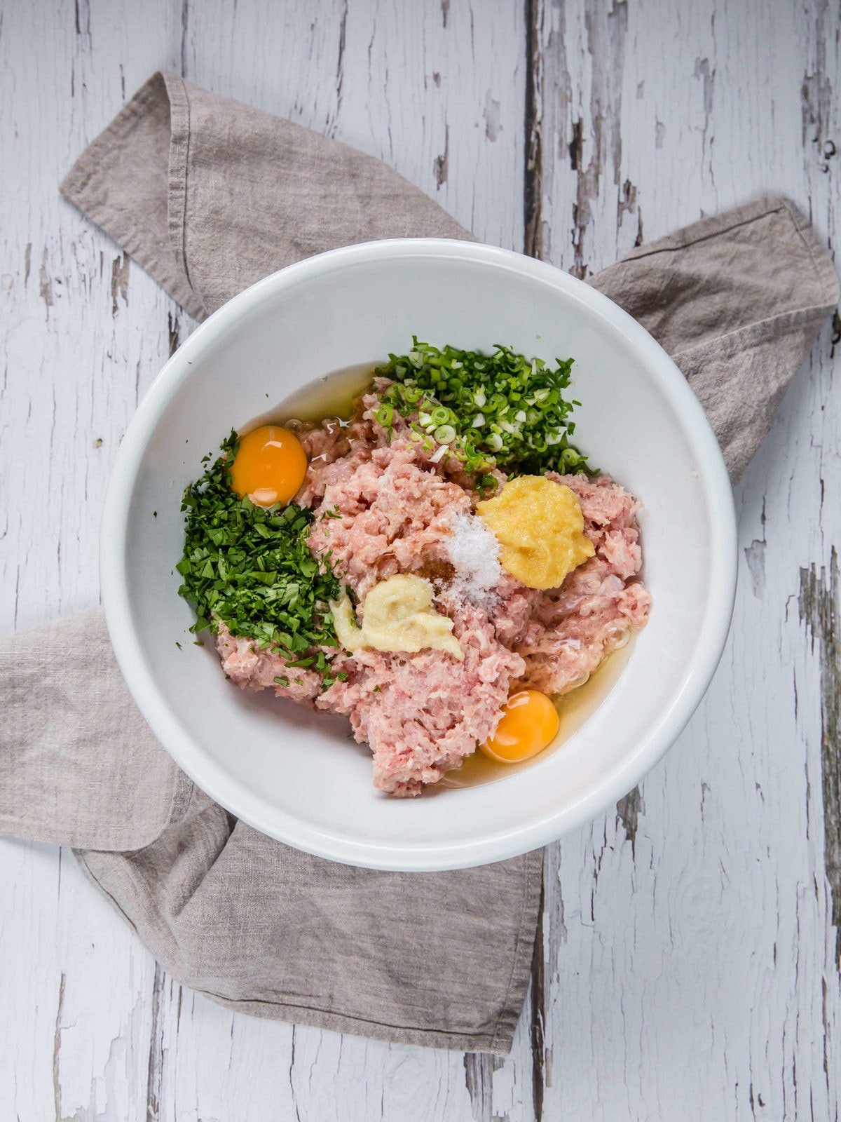 a white mixing bowl with ground chicken, ginger, garlic, egg, cilantro, green onion, sesame oil and coconut flour 