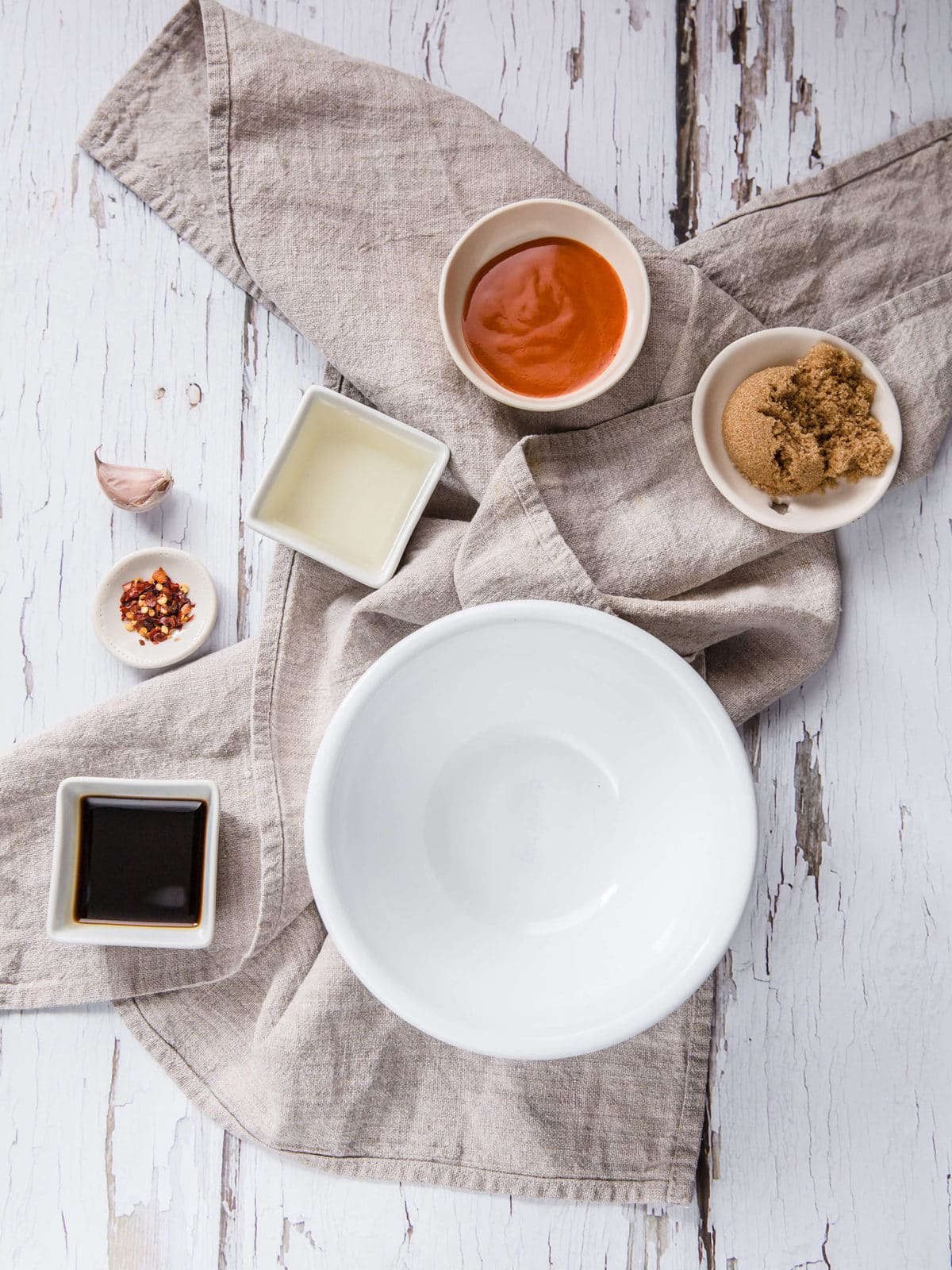 a white bowl, surround by ingredients to make an asian dipping sauce - soy sauce, vinegar, pepper flakes, hot sauce, sweetener, and garlic