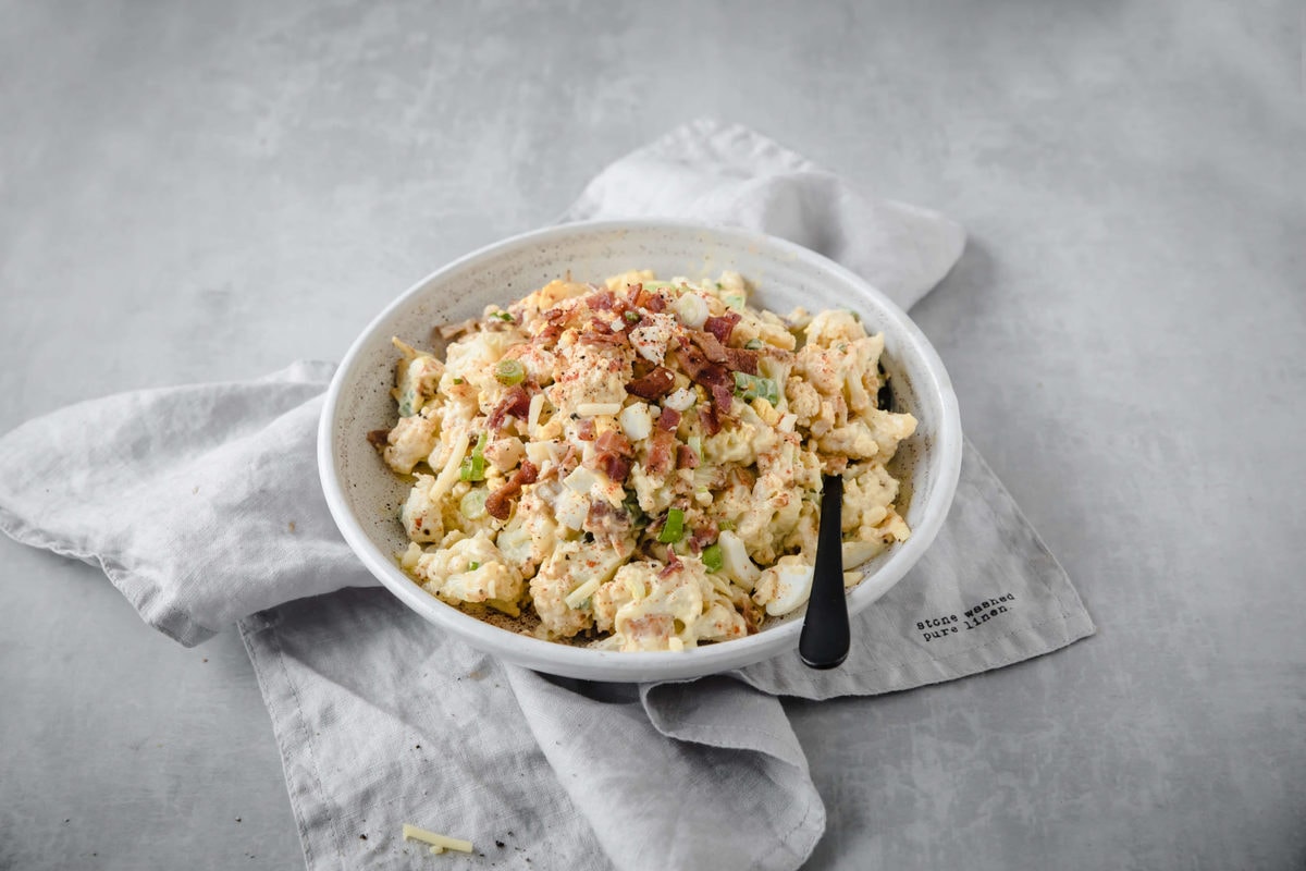 a white bowl full of keto potato salad, with a black serving spoon and gray dish towel