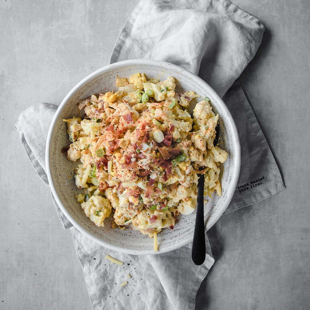 a white bowl with a loaded cauliflower salad with eggs, olives, green onions bacon and black pepper on top