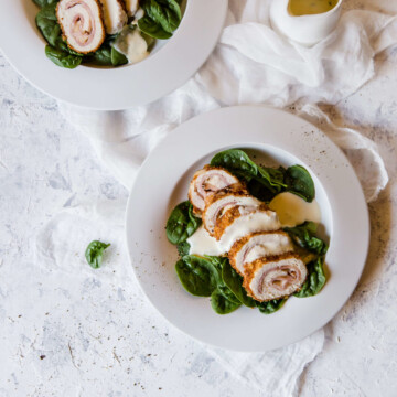 Two white plates of sliced medallions of Keto Chicken Cordon Bleu on a bed of raw spinach, topped with creamy cheese sauce. A white cream saucer with extra creamy cheese sauce sits to the side.