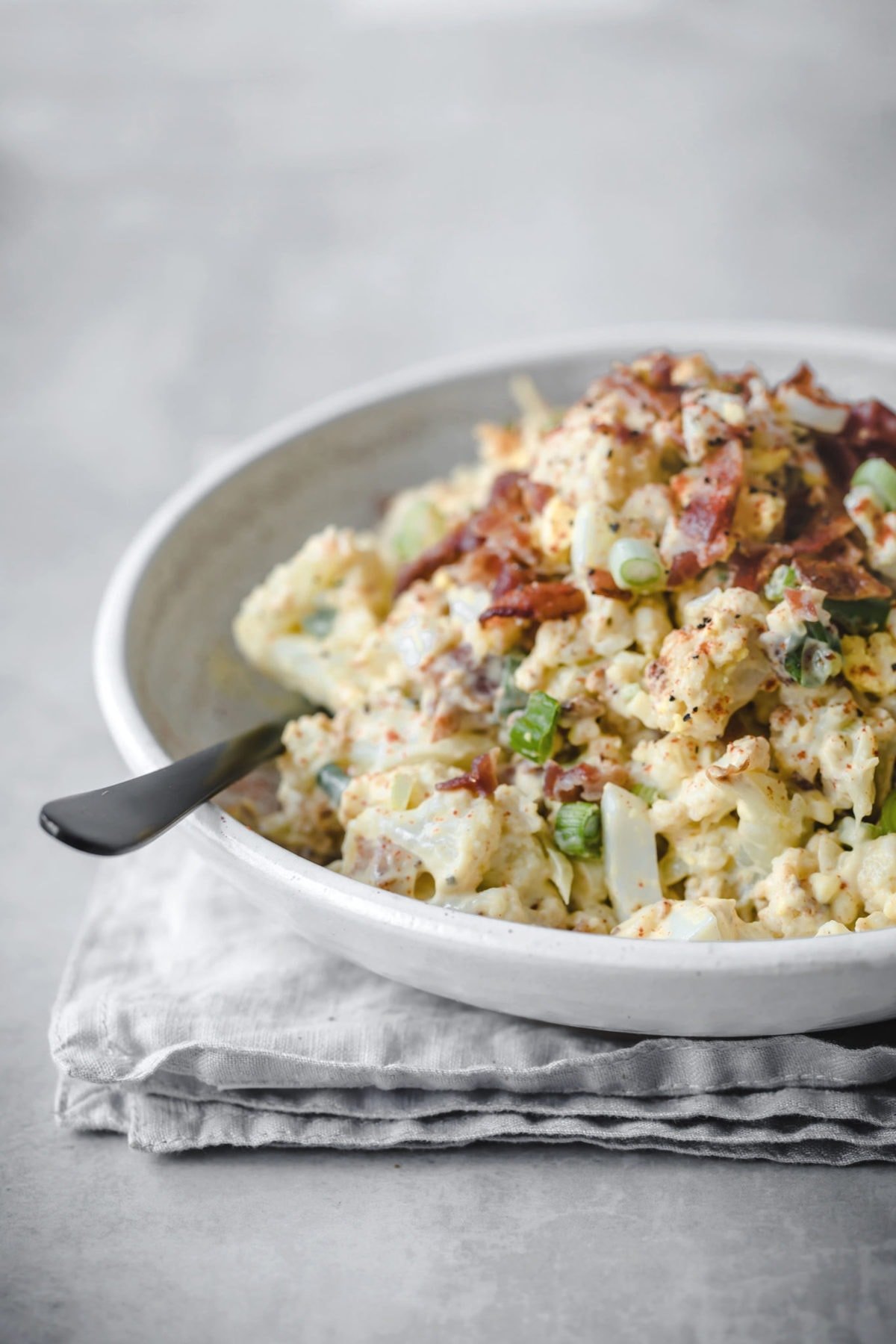 a white bowl with a loaded cauliflower salad with eggs, olives, green onions bacon and black pepper on top