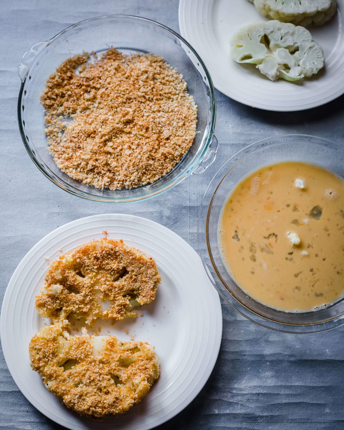 a white plate with breaded cauliflower steaks on it. With a bowl of egg wash and a bowl of breading next to it.