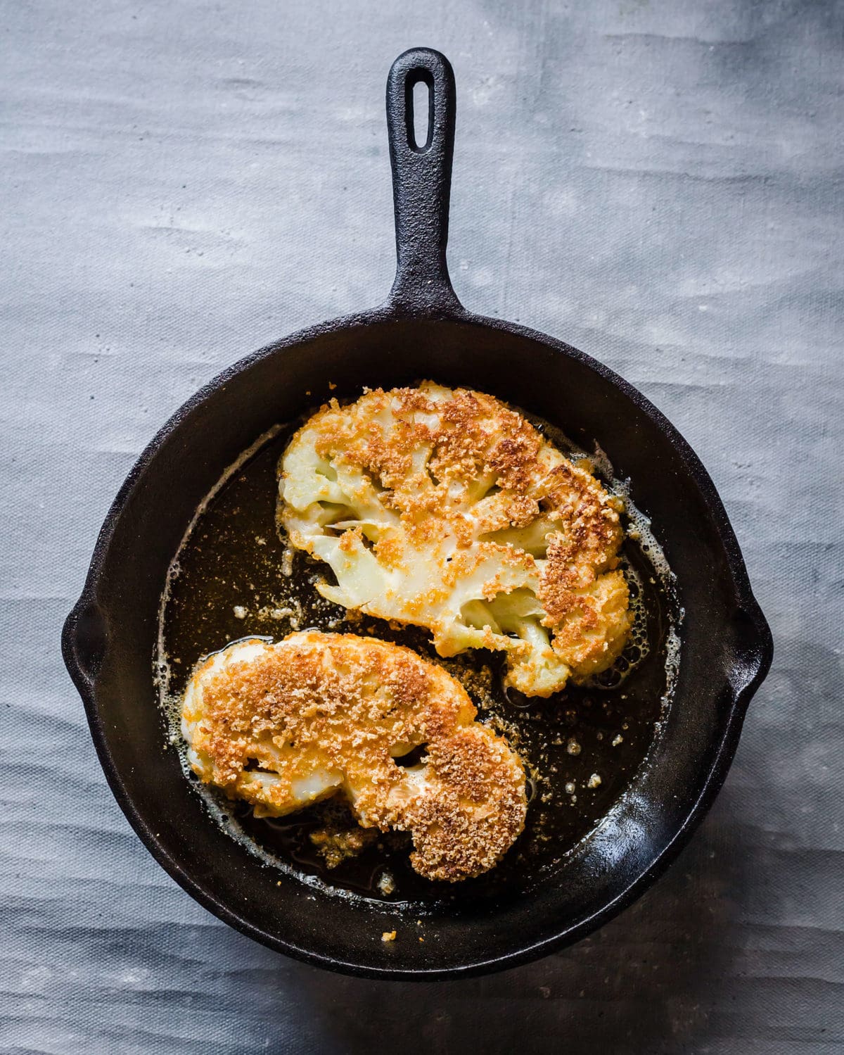 a cast iron skillet with 2 breaded cauliflower steaks being fried until crispy. 