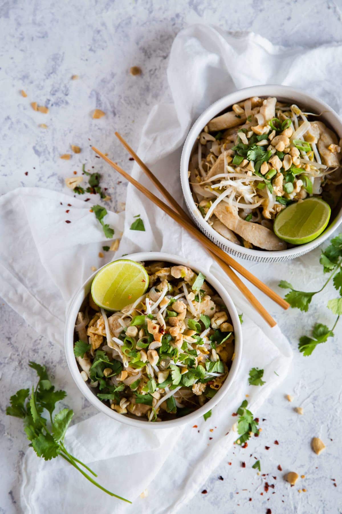 2 white bowls, full of keto Pad Thai, garnished with green onion, peanuts, cilantro and lime wedges and served with chopsticks