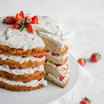 Keto Strawberry Shortcake with 4 layers of cake, whipped cream, and strawberries. The cake sits on a white cake stand and a piece is cut out. Strawberries are scattered on the white tablecloth in the background.