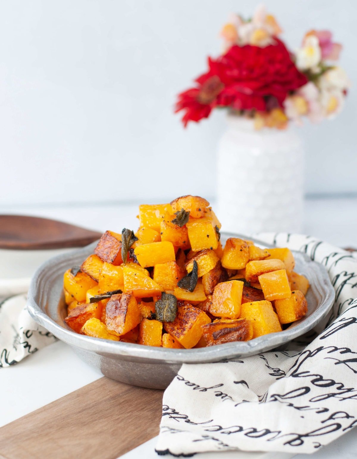 A silver serving dish full of cubed up, roasted butternut squash, garnished with crispy sage and fresh sage on the side. 