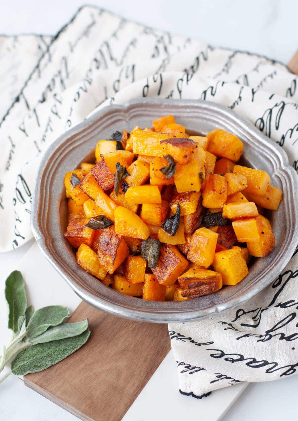 A silver serving dish full of cubed up, roasted butternut squash, garnished with crispy sage and fresh sage on the side. 