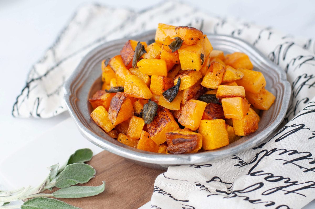 A silver serving dish full of cubed up, roasted butternut squash, garnished with crispy sage and fresh sage on the side. 