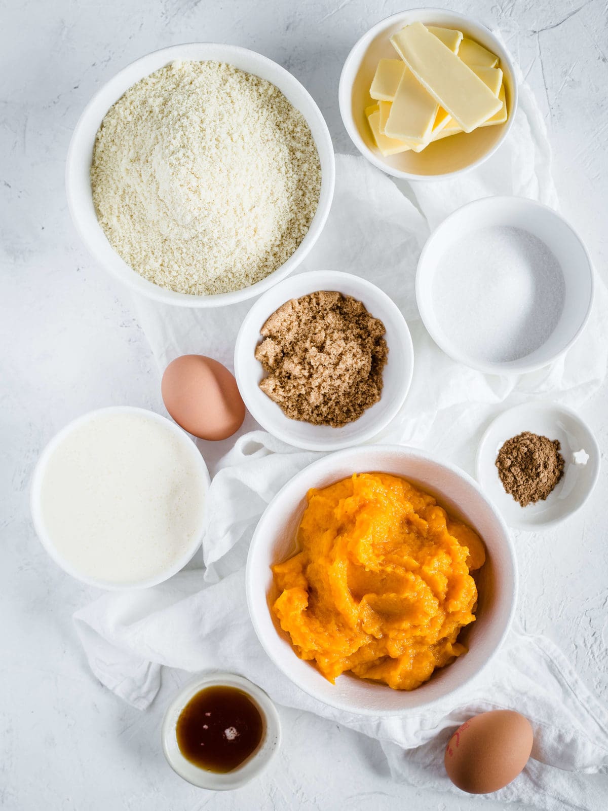 Ingredients laid out in individual bowls to make a low carb pumpkin pie - almond flour, eggs, butter, sweeteners, pumpkin pie spice, pumpkin puree and vanilla.