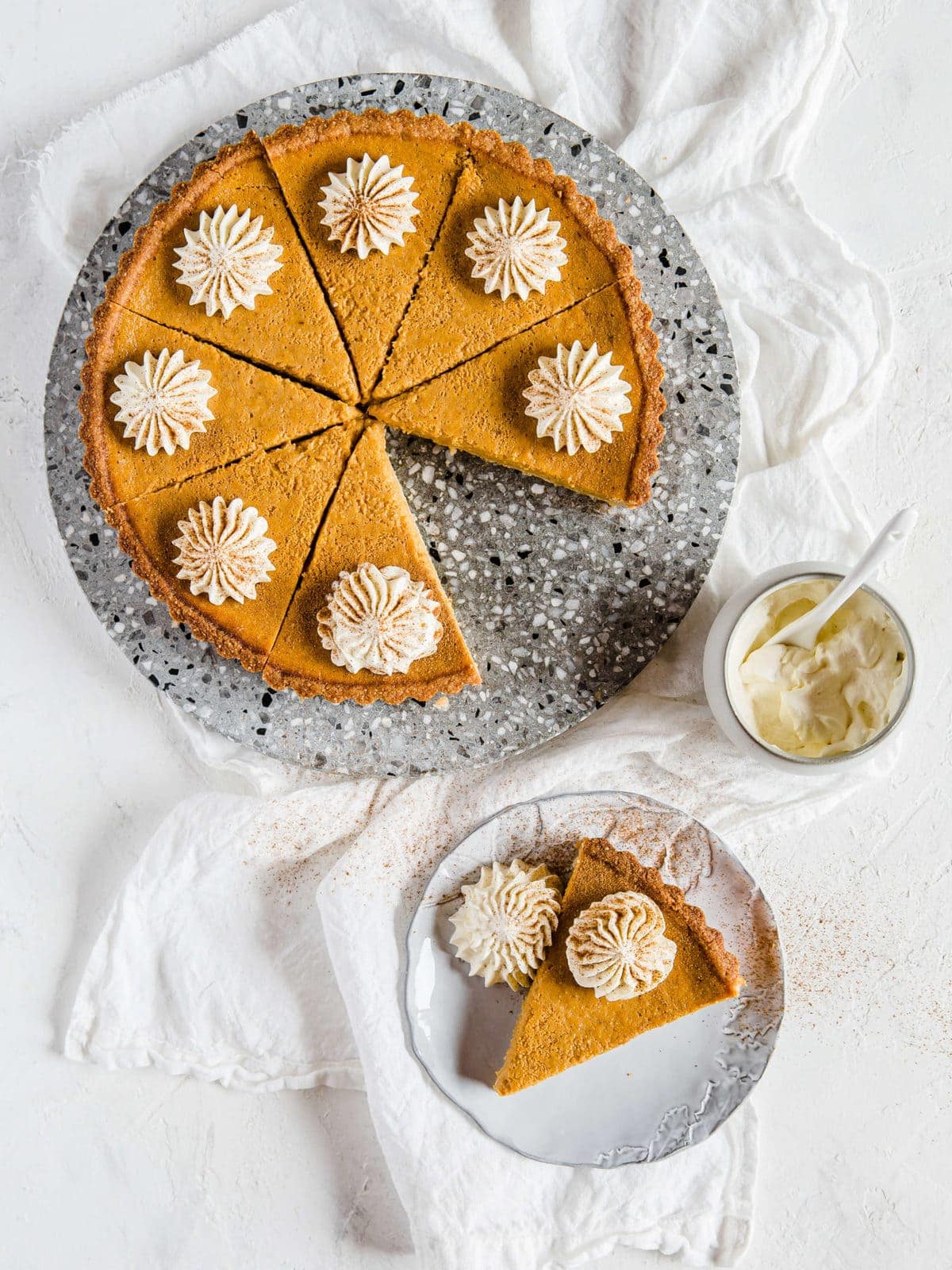 overhead shot of a pumpkin pie on a platter, with a plate next to it with a single slice, topped with fresh whipped cream.
