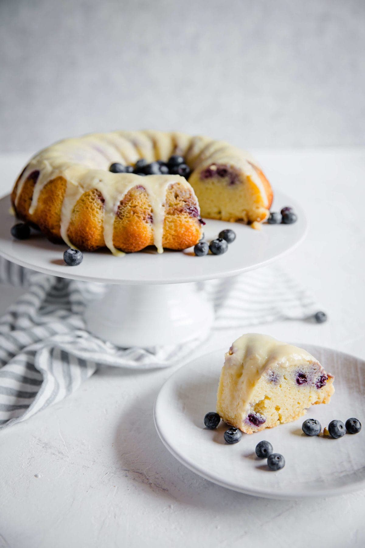 Low carb blueberry lemon pound cake, with lemon glaze and fresh blueberries on a cake stand, with a plated piece of cake in front of it. 