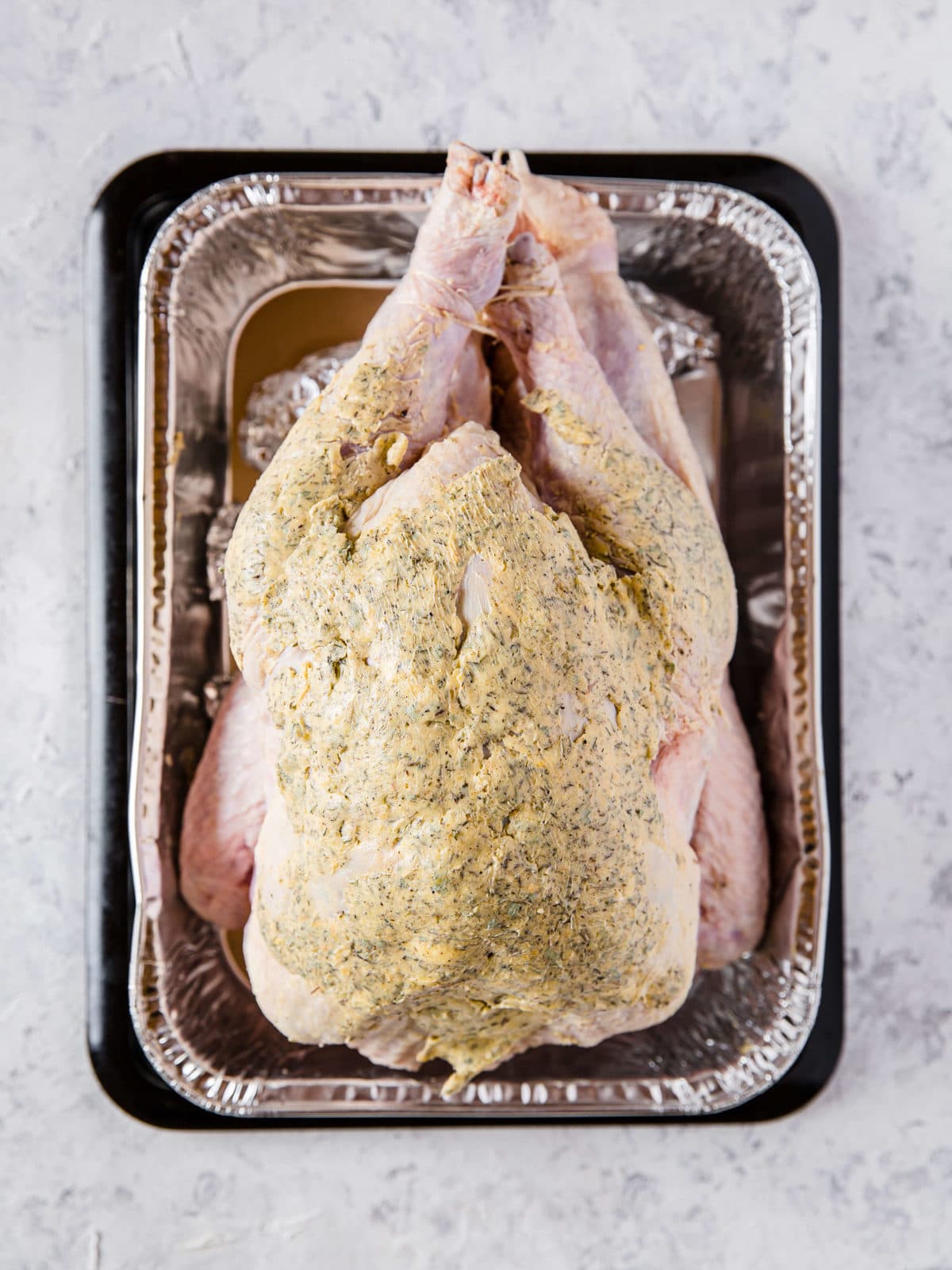 An aluminum roasting pan with a raw turkey in it that is covered with compound butter.