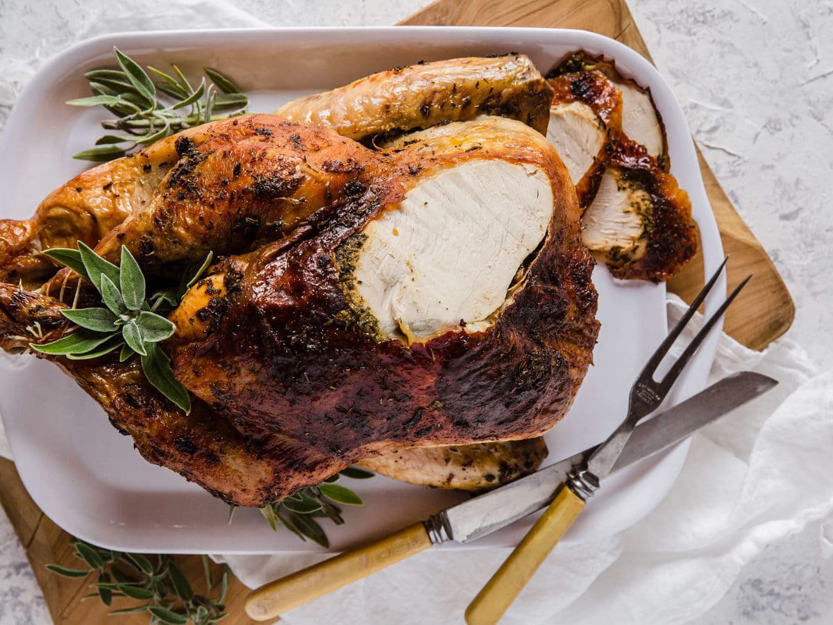 An Herb Butter Roasted Turkey fresh out of the oven, surrounded by fresh herbs and carving utensils.