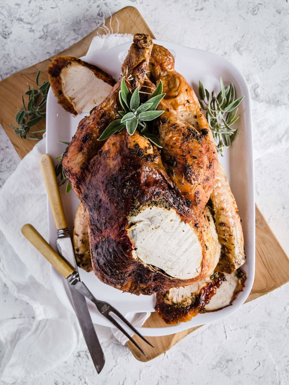 An Herb Butter Roasted Turkey fresh out of the oven, surrounded by fresh herbs and carving utensils.