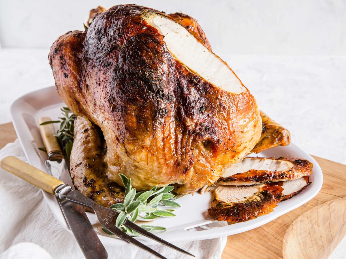 An Herb Butter Roasted Turkey fresh out of the oven, surrounded by fresh herbs and carving utensils.