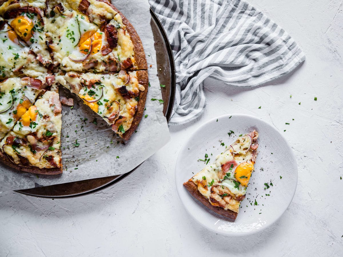 overhead shot of a low carb pizza crust, topped with cream sauce and bacon, cheese, and eggs