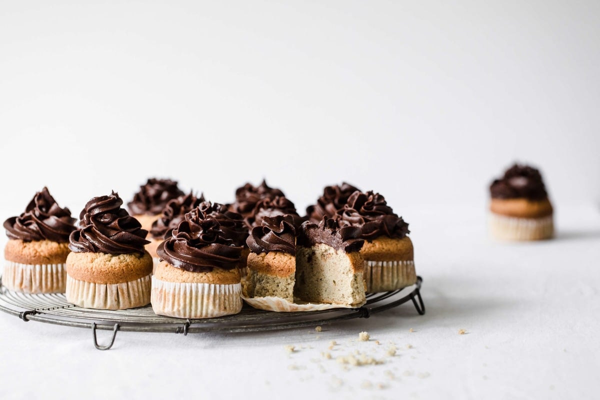 vanilla cupcakes with chocolate frosting featured on a cooling rack with one cupcake missing a slice