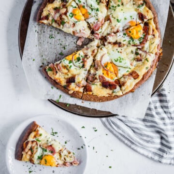 Keto Breakfast Pizza, cut into slices, sitting on a sheet of white parchment paper on a pizza stone. A single slice of breakfast pizza sits on a small white plate.