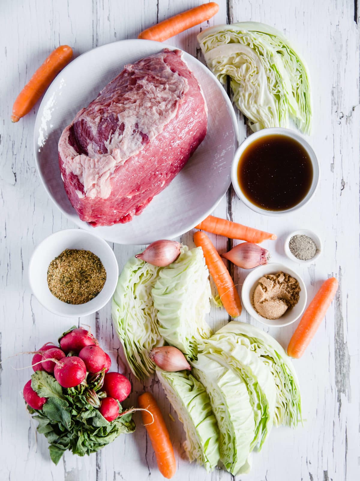 An overhead shot of all of the ingredients to make corned beef and cabbage: beef brisket with the excess fat removed, beef broth, corned beef seasonings, brown sugar erythritol, radishes, green cabbage, shallots, and carrots.