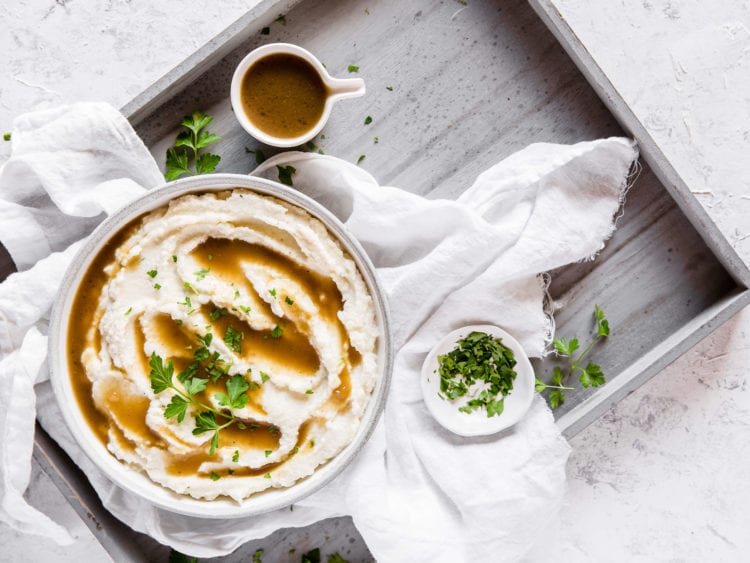 An overhead shot of a bowl of creamy cauliflower mash, topped with keto pan gravy and garnished with fresh herbs, with a creamer dish of extra gravy and a small bowl of fresh herbs to the side.