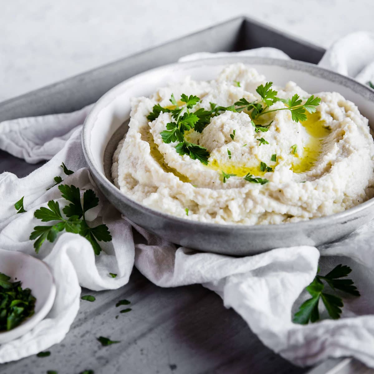 a gray ceramic bowl full of mashed cauliflower with a swirl of melted butter and parsley on top