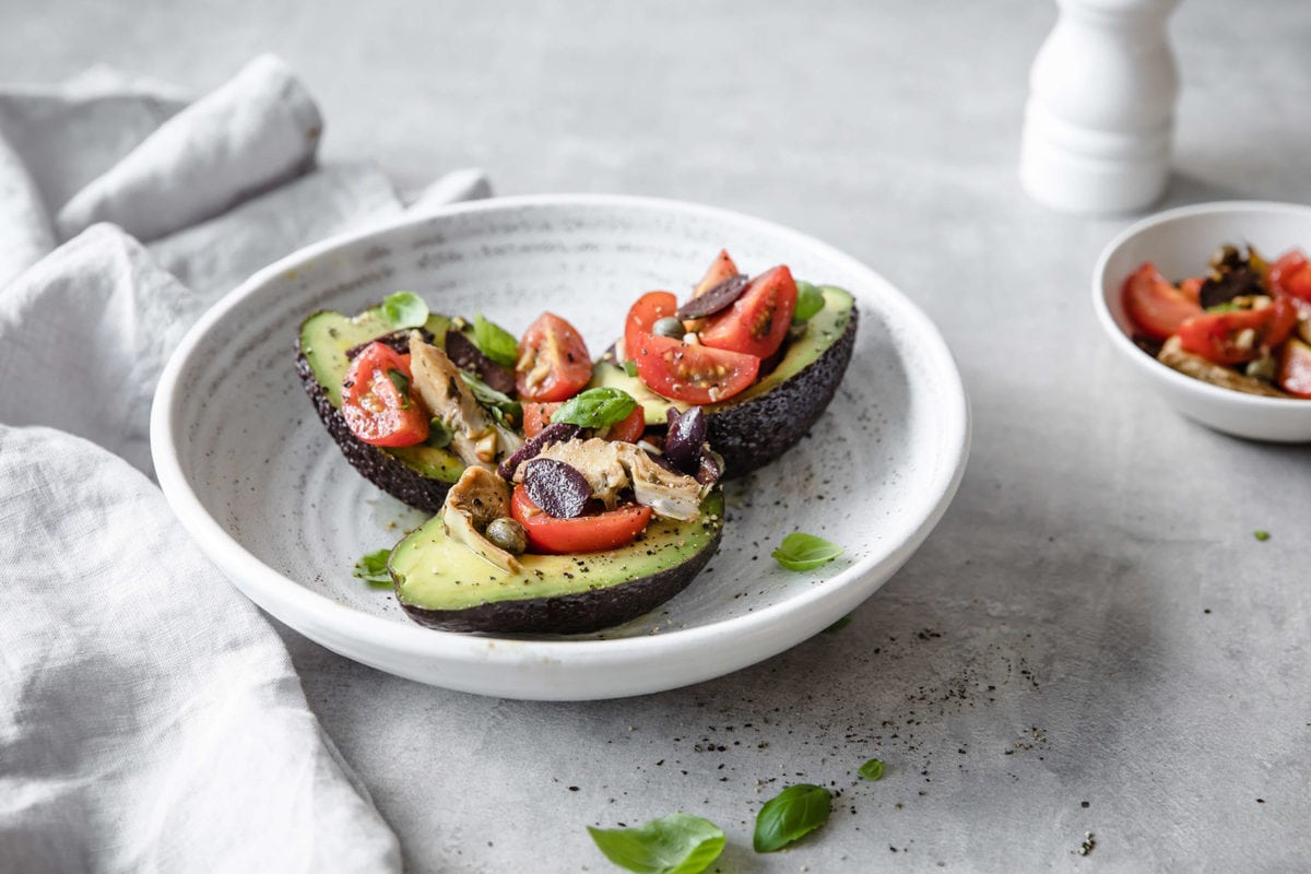 Three avocado halves, sliced lengthwise with the seed removed, stuffed with bruschetta, served on a round, rustic white, ceramic plate, garnished with basil leaves.