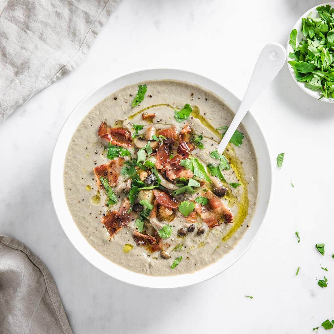 Creamy mushroom soup garnished with a swirl of olive oil, crispy bacon, fresh parsley, and roasted mushrooms and garlic