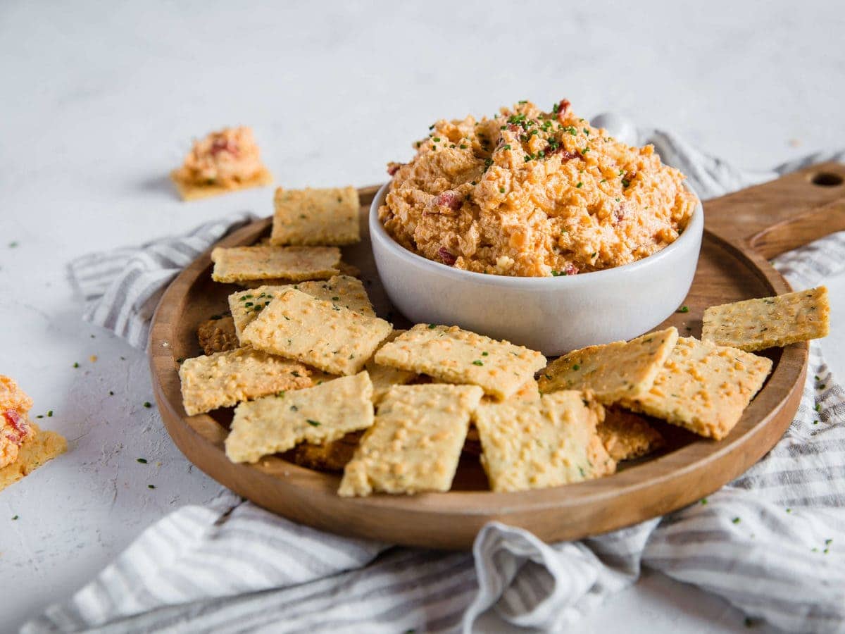 A head on shot of a cheese dip dish overflowing with Pimento Cheese Dip, sitting on a wooden charcuterie board piled with Parmesan Chive and Garlic Keto Crackers.