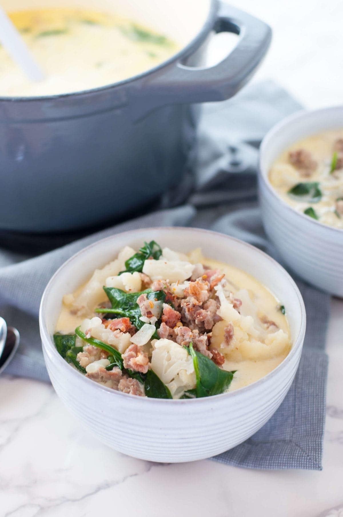 Overhead shot of a white soup bowl filled to the brim with creamy keto Zuppa Toscana, topped with chopped crispy bacon and parmesan cheese, on a gray napkin, sitting on top of a marble countertop. In the background, there is a gray blue dutch oven full of soup, plus a second bowl and silver spoons, ready to dig in.