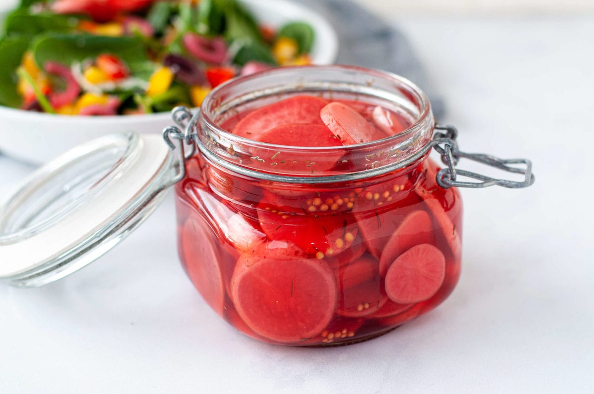 Side view of a small glass jar with a clamp lid, full of bright red quick pickled radishes, sitting on a marble countertop. In the background, there's a large white bowl filled with salad, waiting to be topped with picked radishes.