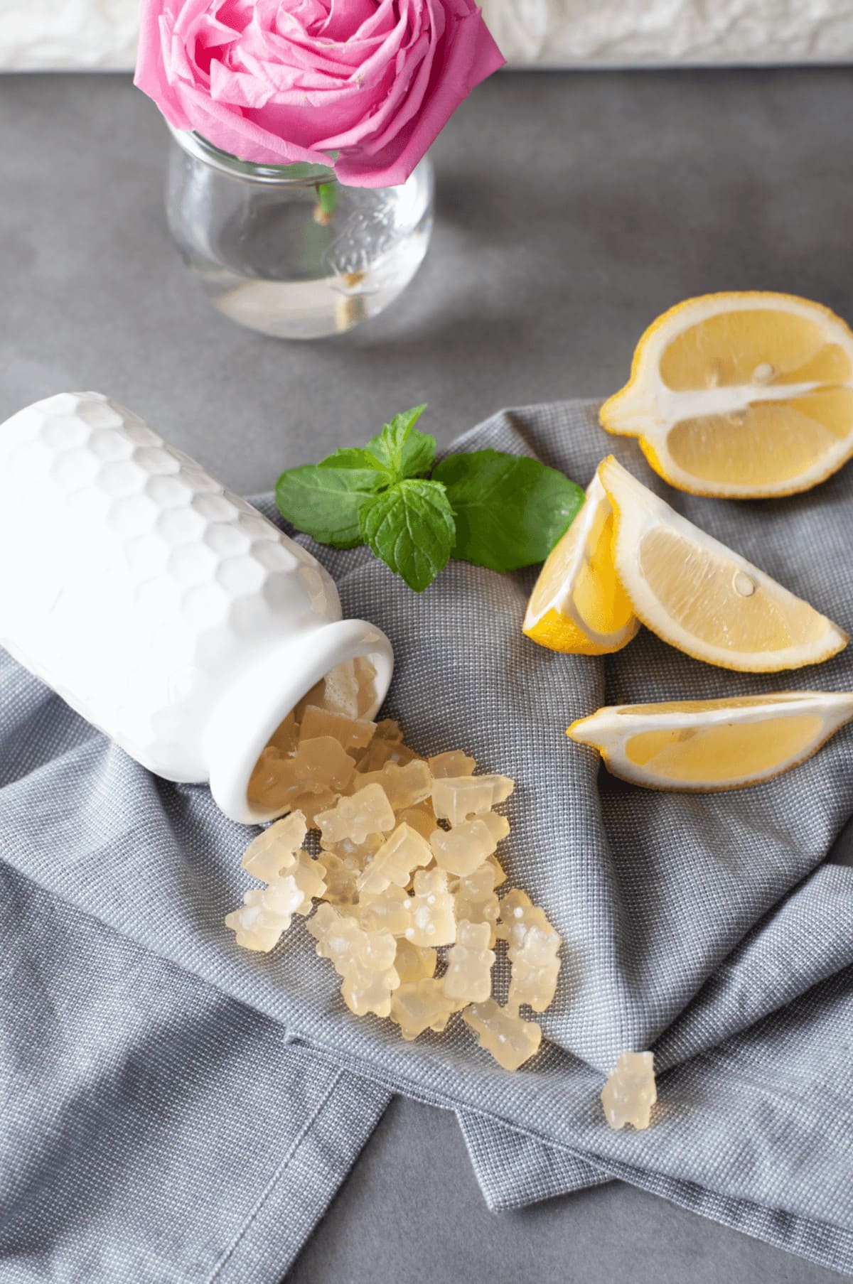 Fresh mint and lemons next to a white jar, spilling out lemon gummy bears onto a gray napkin. 