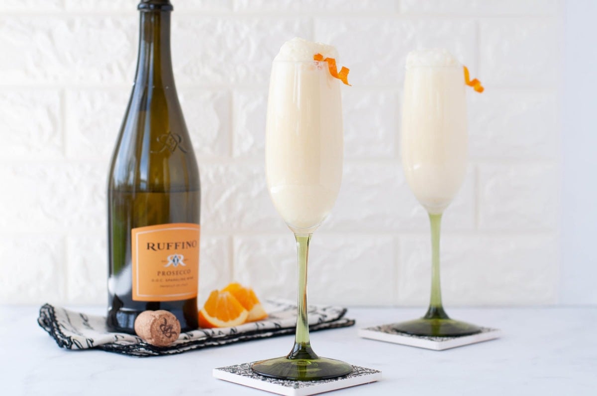 two champagne flutes full of a creamy mimosa, garnished with a twist of orange and a bottle of wine in the background 