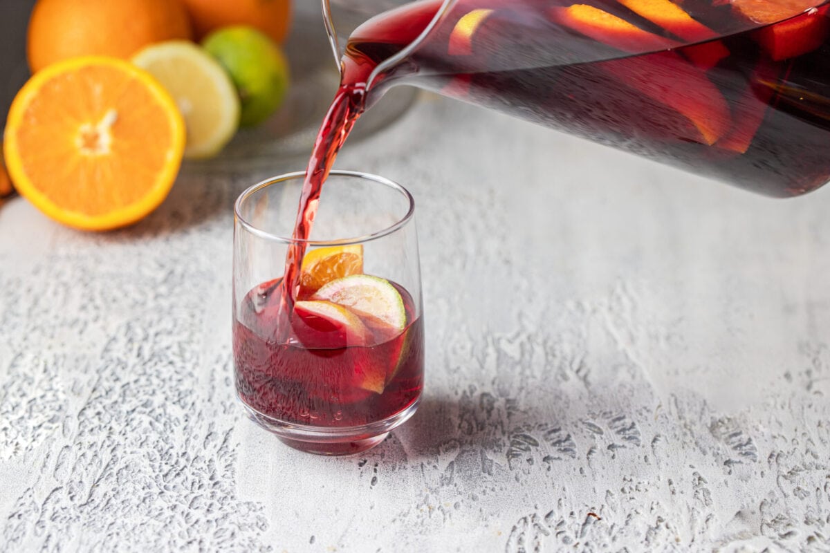 A glass of kombucha being poured from a pitcher. Garnished with limes, lemons and oranges 