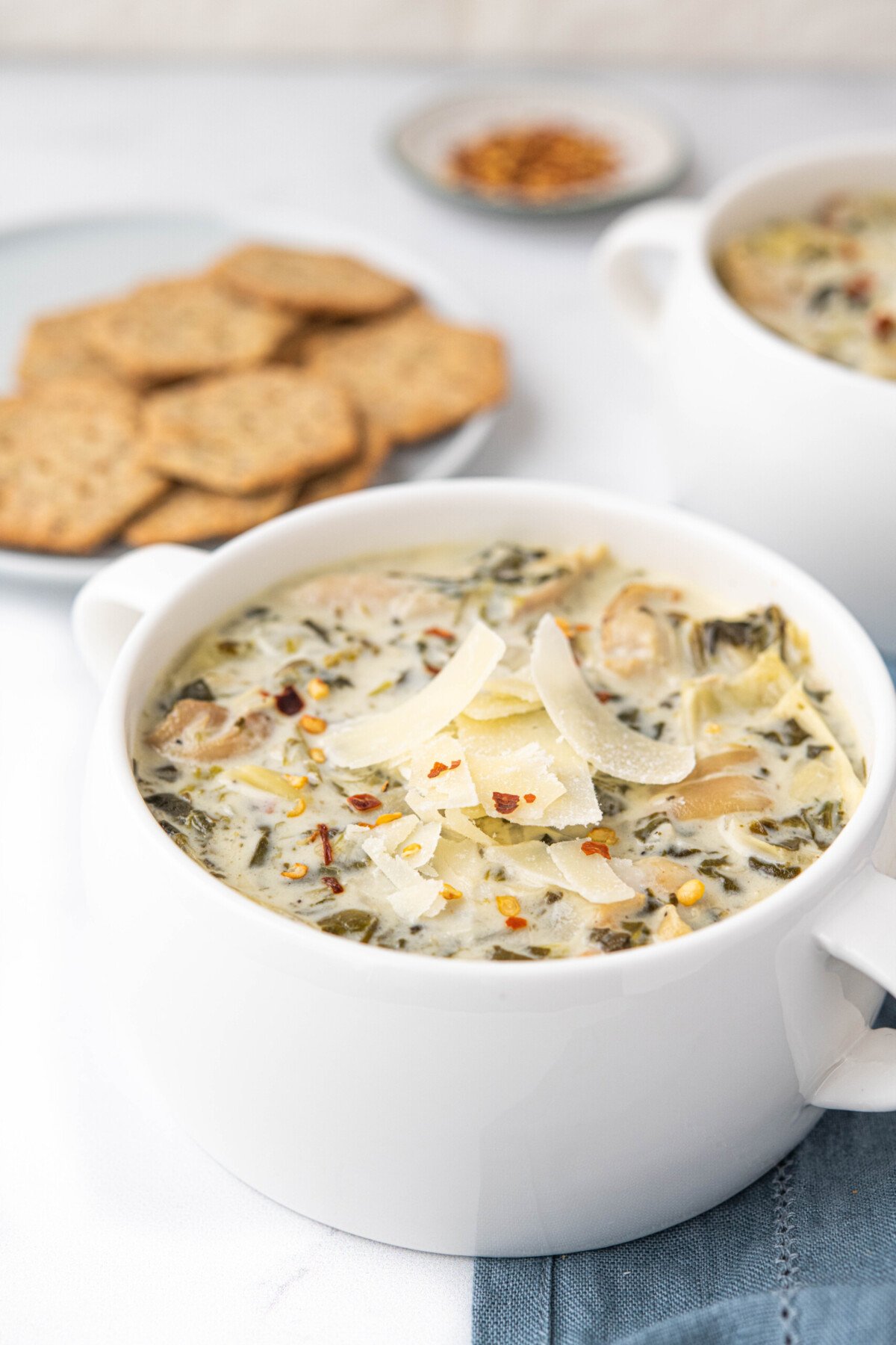 Bowl of spinach and artichoke soup in a white bowl, topped with shaved parmesan cheese and red pepper flakes