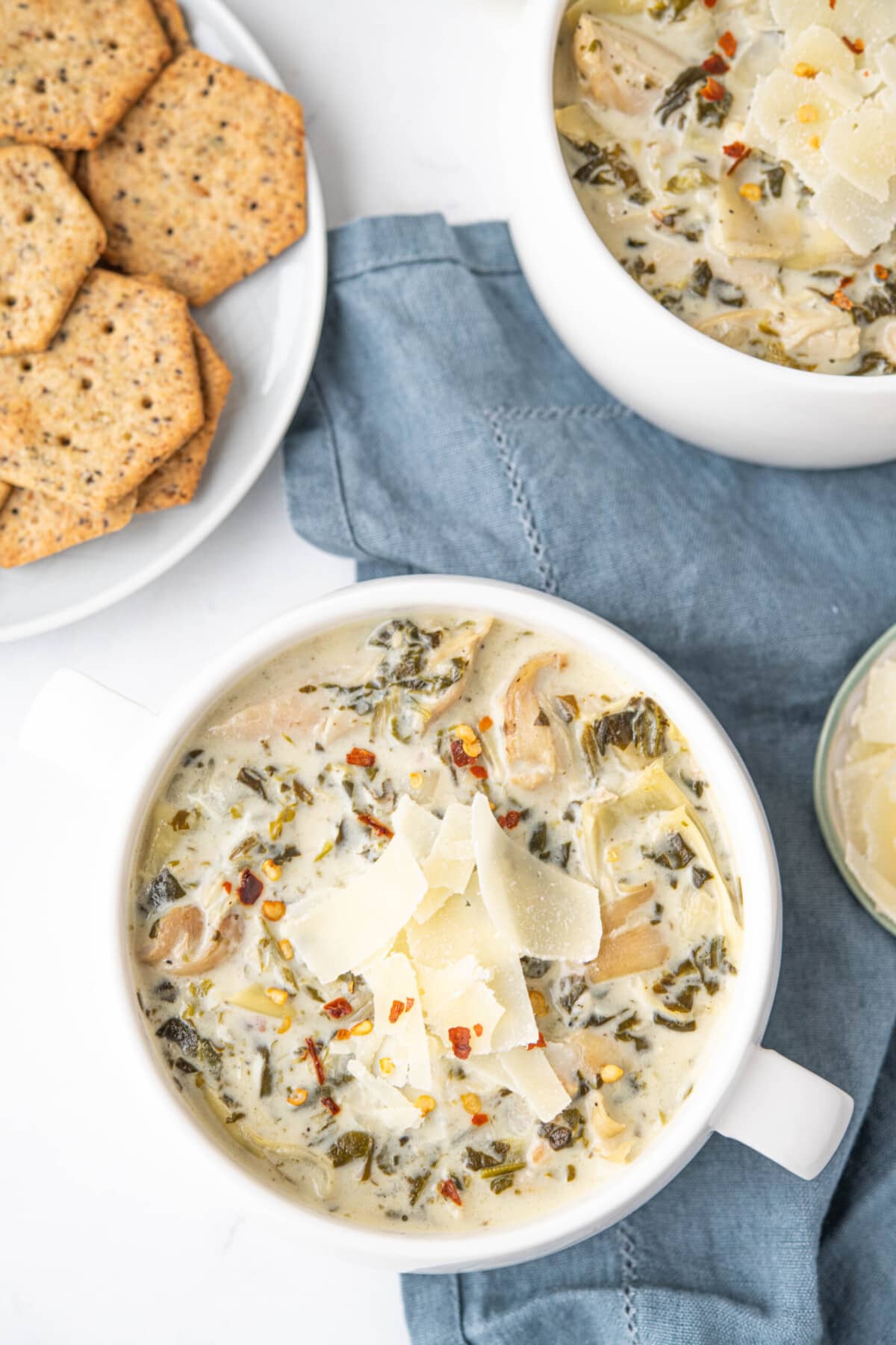 Bowl of spinach and artichoke soup in a white bowl, topped with shaved parmesan cheese and red pepper flakes.