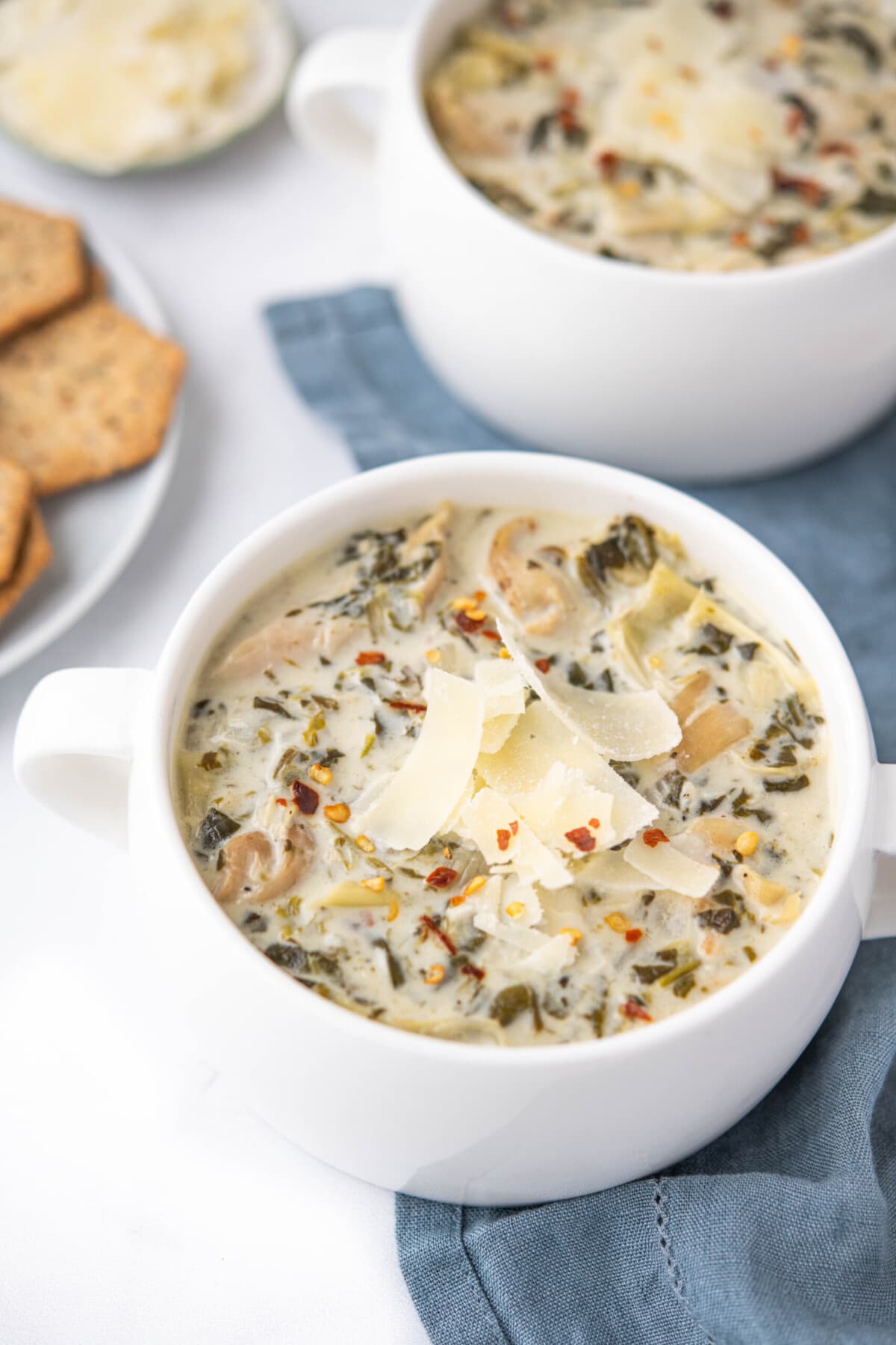 Bowl of spinach and artichoke soup in a white bowl, topped with shaved parmesan cheese and red pepper flakes.