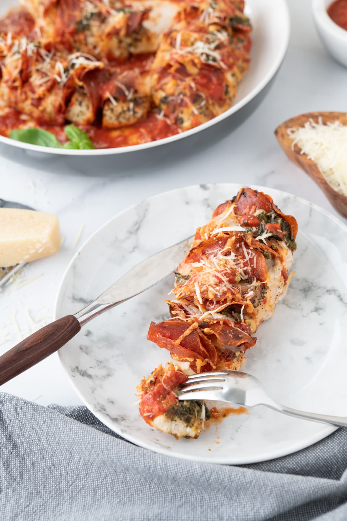 Chicken breast with pepperoni, pizza sauce and cheese, garnished with cheese and pesto, on a marble plate with a fork and knife