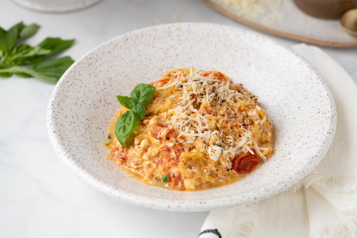 A specked pottery bowl filled with a low carb baked feta pasta with tomatoes, basil and topped with cheese