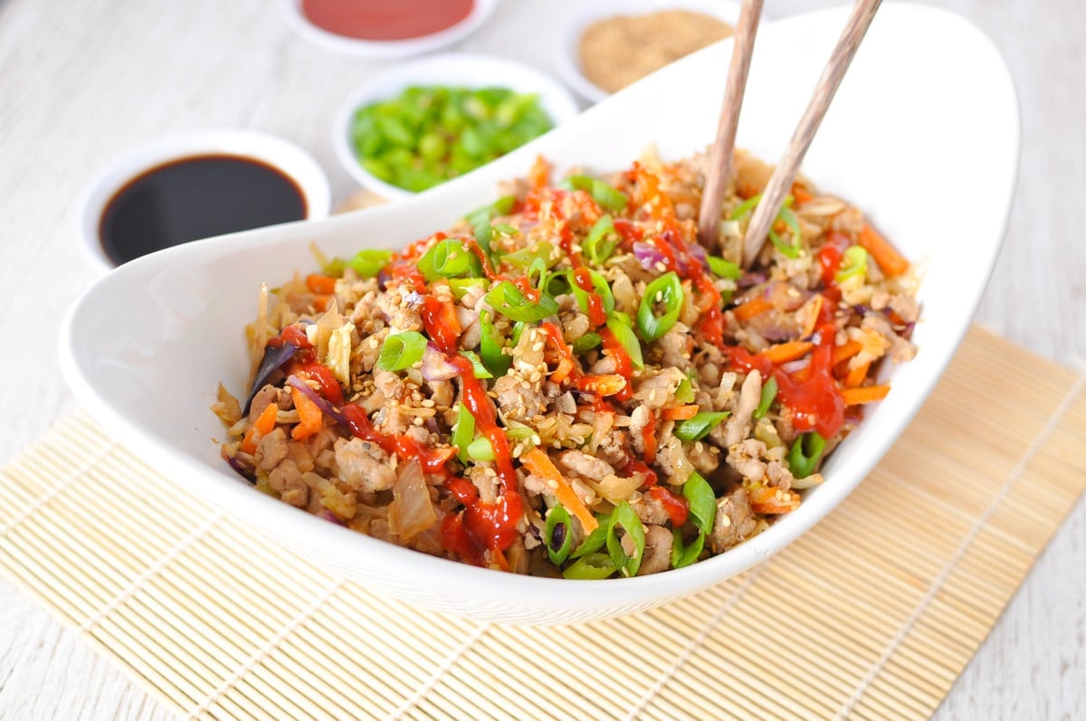 A white serving dish filled with egg roll in a bowl, served with chopsticks and garnished with green onions and sriracha