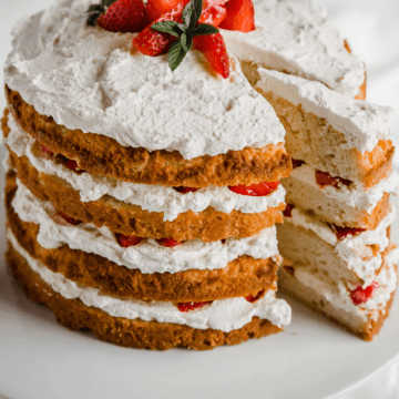 Keto Strawberry Shortcake with 4 layers of cake, whipped cream, and strawberries. The cake sits on a white cake stand and a piece is cut out.