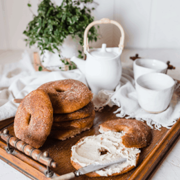 Cinnamon sugar(free) bagels are stacked on a wooden tray with a white tea set and a potted plant in the background. One bag is cut in half and slathered with cream cheese.