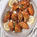 Metal serving dish with crispy chicken wings, topped with feta cheese and parsley, and garnished with lemon