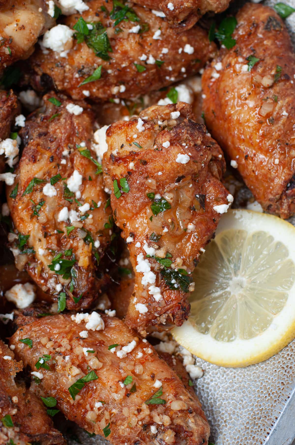 Metal serving dish with crispy chicken wings, topped with feta cheese and parsley, and garnished with lemon
